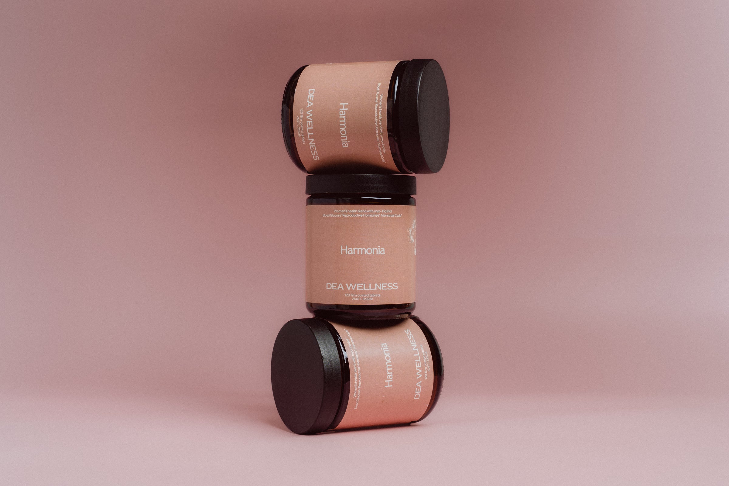 Three amber glass jars with black lids stacked vertically on a soft pink background. The labels are a warm peach-beige color with white text reading 'Harmonia' and 'DEA WELLNESS'.
