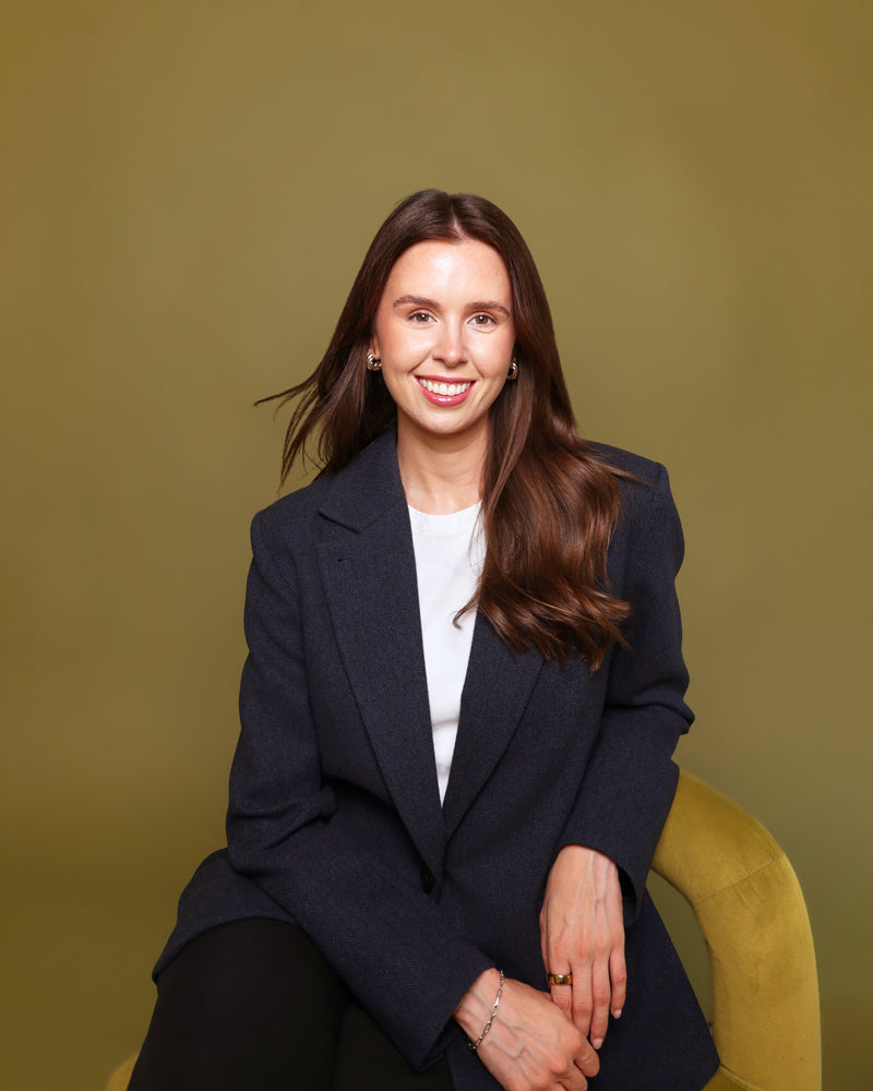 A smiling woman with long brown hair sits on a mustard yellow chair against a solid olive-green studio background. She is wearing a dark navy blazer over a white top and black pants.
