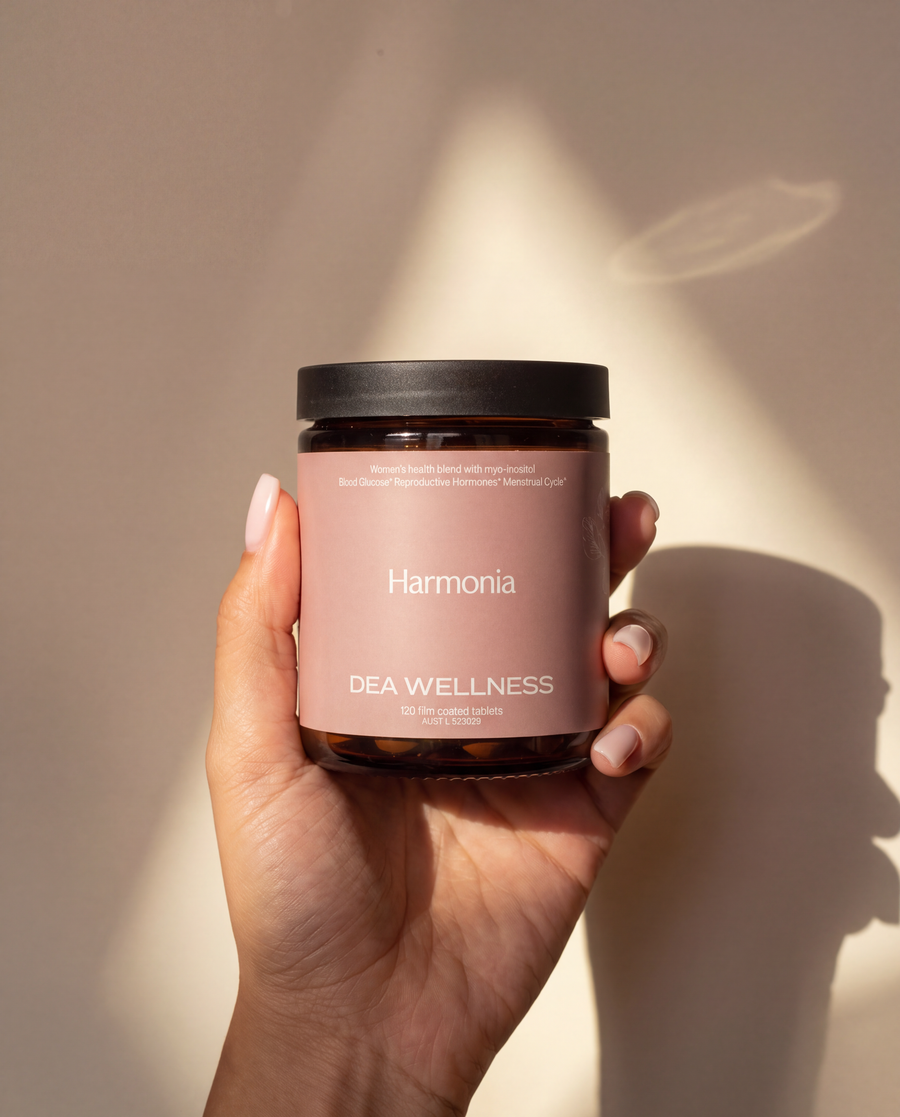 A hand holding a amber glass jar candle with a dusty pink label and a black lid. The label reads 'Harmonia' and 'DEA WELLNESS'.