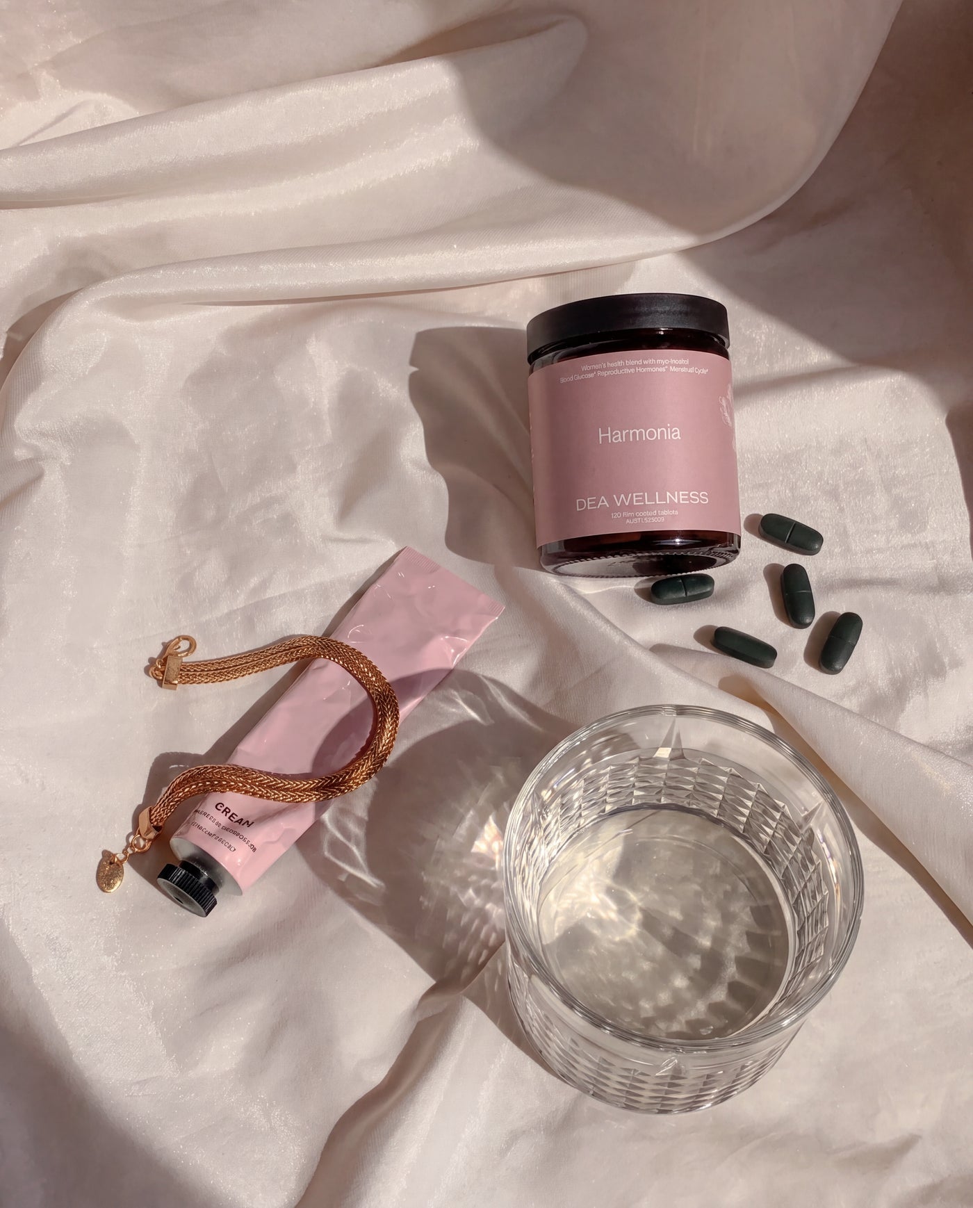 A styled flat-lay of a pink jar labeled 'Harmonia' and 'DEA WELLNESS' alongside a pink tube, a gold chain, dark green capsules, and a clear textured glass on draped white fabric.