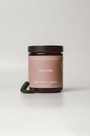 A dark amber glass jar with a dusty pink label reading 'Harmonia' and 'DEA WELLNESS' under it, with a dark green capsule resting beside the base, on a light gray background.