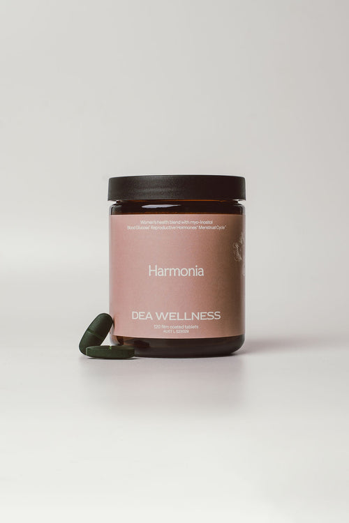 A dark amber glass jar with a dusty pink label reading 'Harmonia' and 'DEA WELLNESS' under it, with a dark green capsule resting beside the base, on a light gray background.