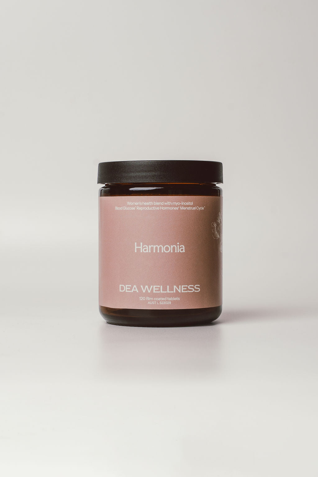A dark amber glass jar with a matte black lid and a dusty pink label reading 'Harmonia' and 'DEA WELLNESS', centered on a light gray background.