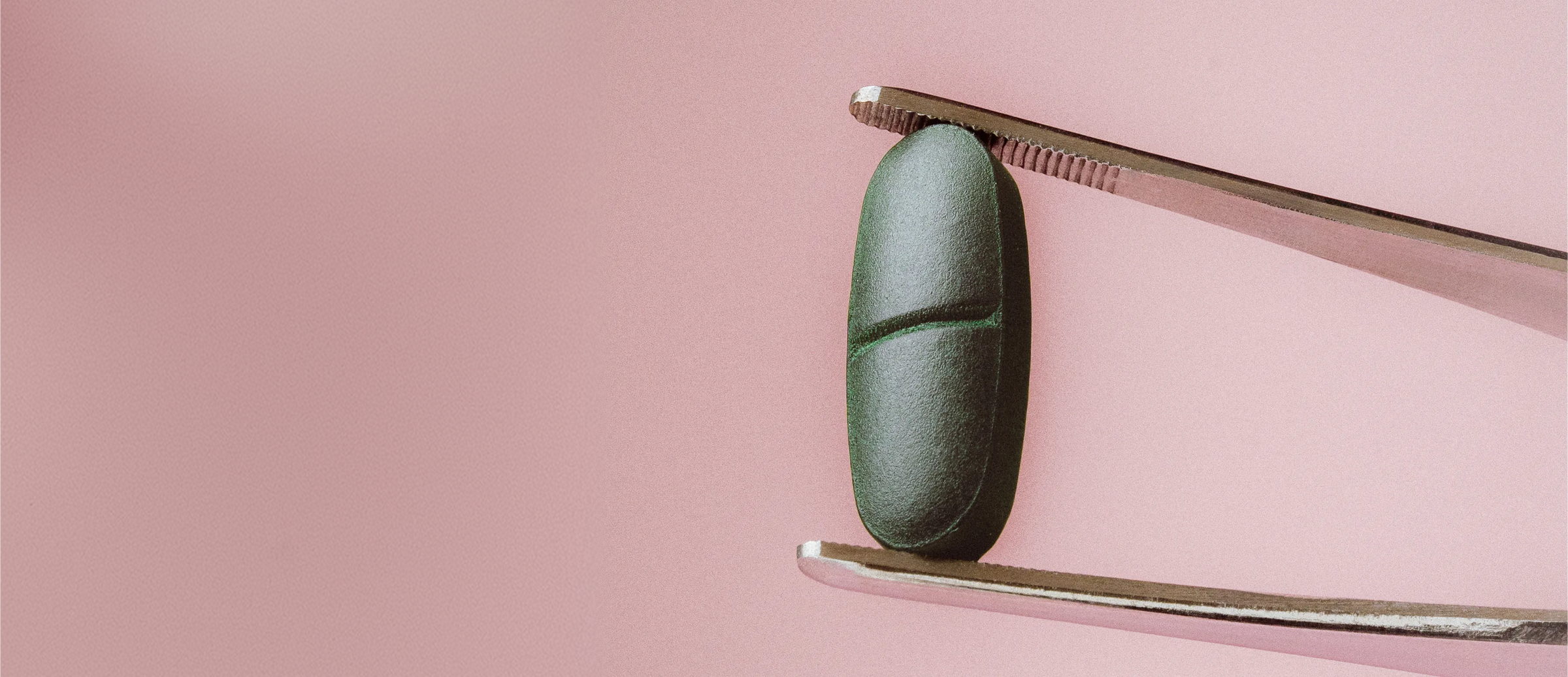 A dark green, oblong capsule pill held between metal forceps against a solid pink background.