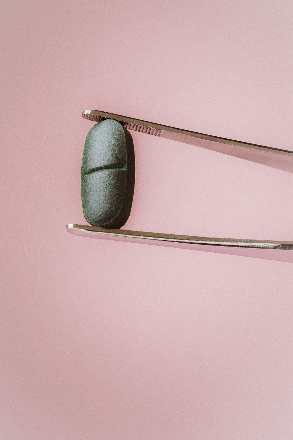 A dark green capsule pill held between silver metal tweezers against a solid pink background.