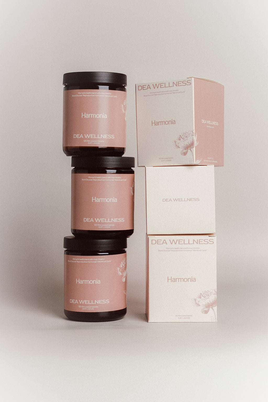 Three amber glass jars with black lids and dusty pink labels are stacked next to three matching product boxes, including two white boxes and one pink box, all branded DEA WELLNESS and Harmonia.