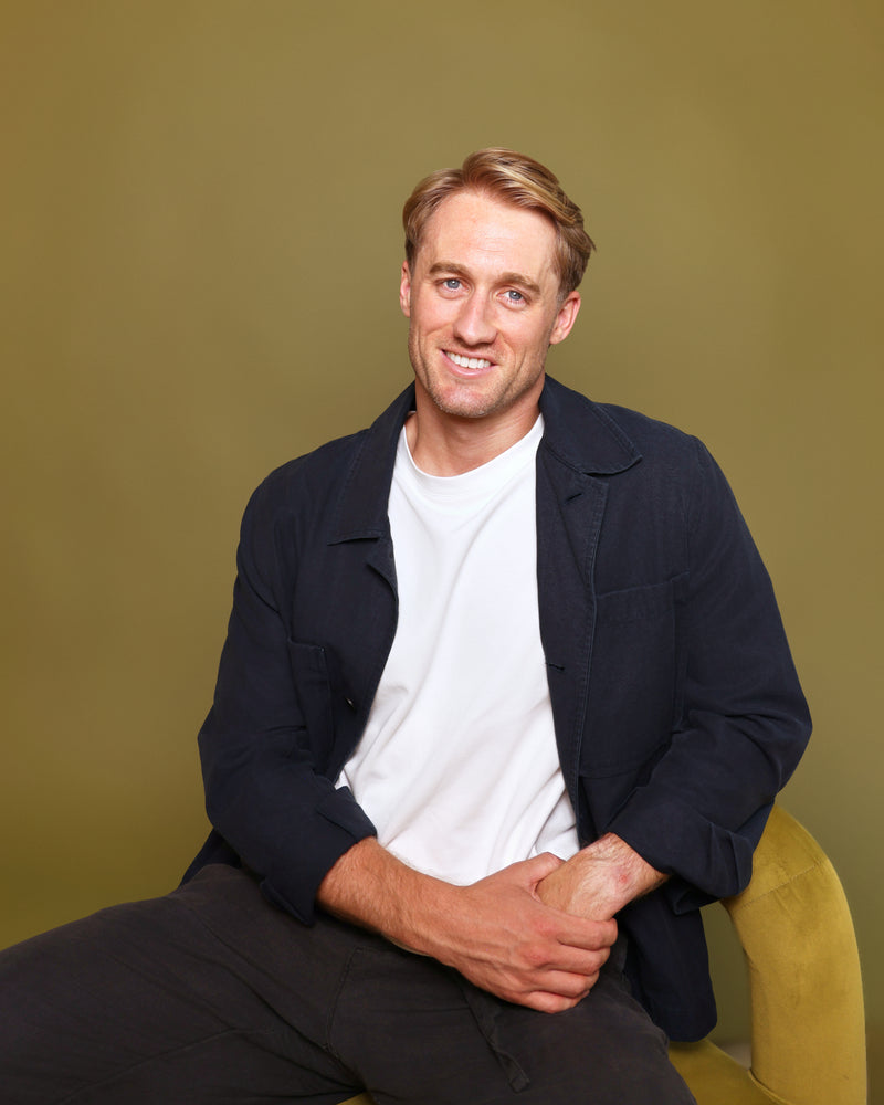 A man with blond hair sitting on a mustard yellow chair against a solid olive-green background. He is wearing a dark navy blue jacket over a white t-shirt and dark trousers.